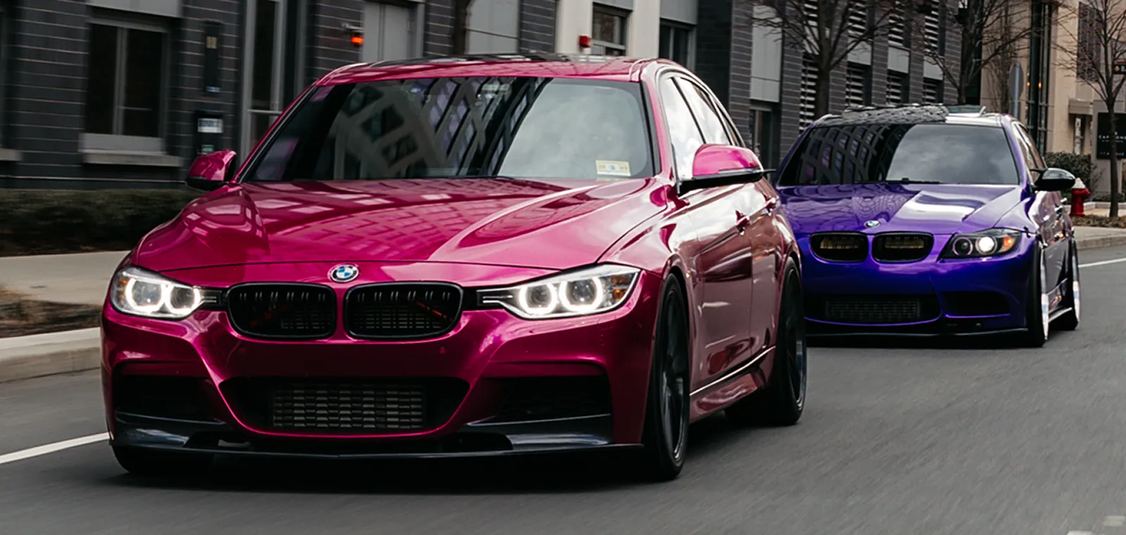 Two BMW cars driving on a city street, one magenta in front and one purple behind.