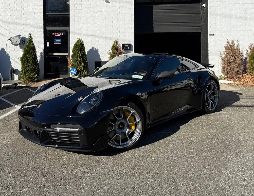 Black Porsche sports car parked on a paved lot in front of a white brick building with a closed door and green shrubs.