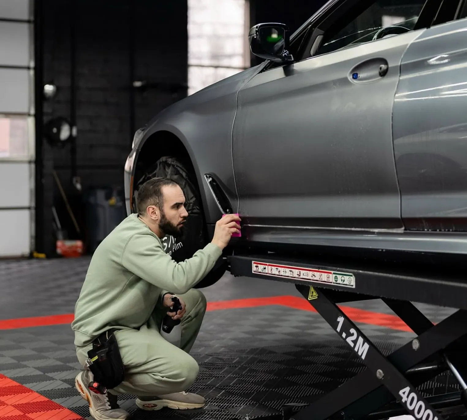 Man in green outfit applying a film or wrap to the side of a gray car on a lift inside a garage.