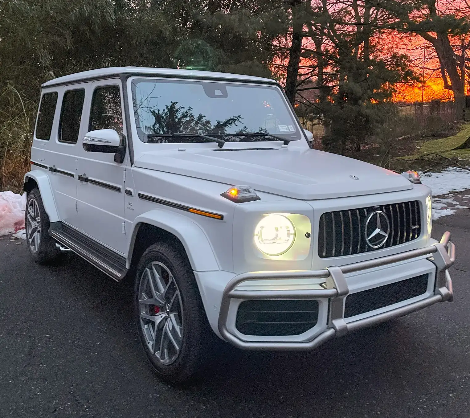 White Mercedes-Benz G-Class SUV with headlights on parked on a road at sunset.