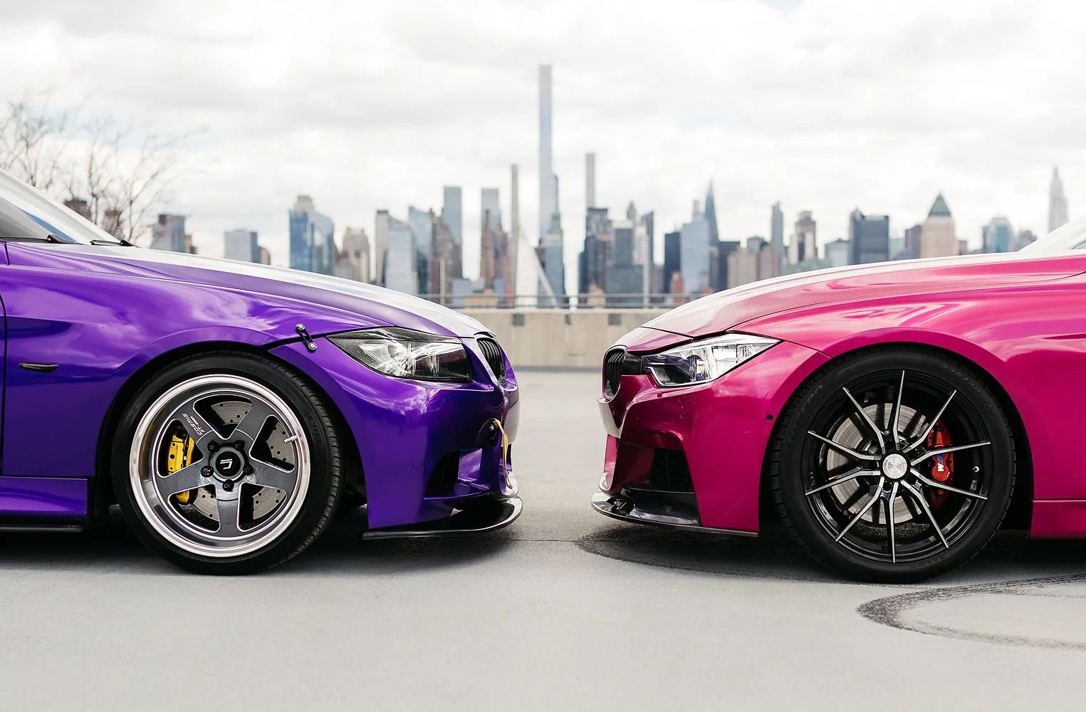 Close-up of the front sides of two modified cars, one purple and one magenta, parked facing each other with a city skyline in the background.