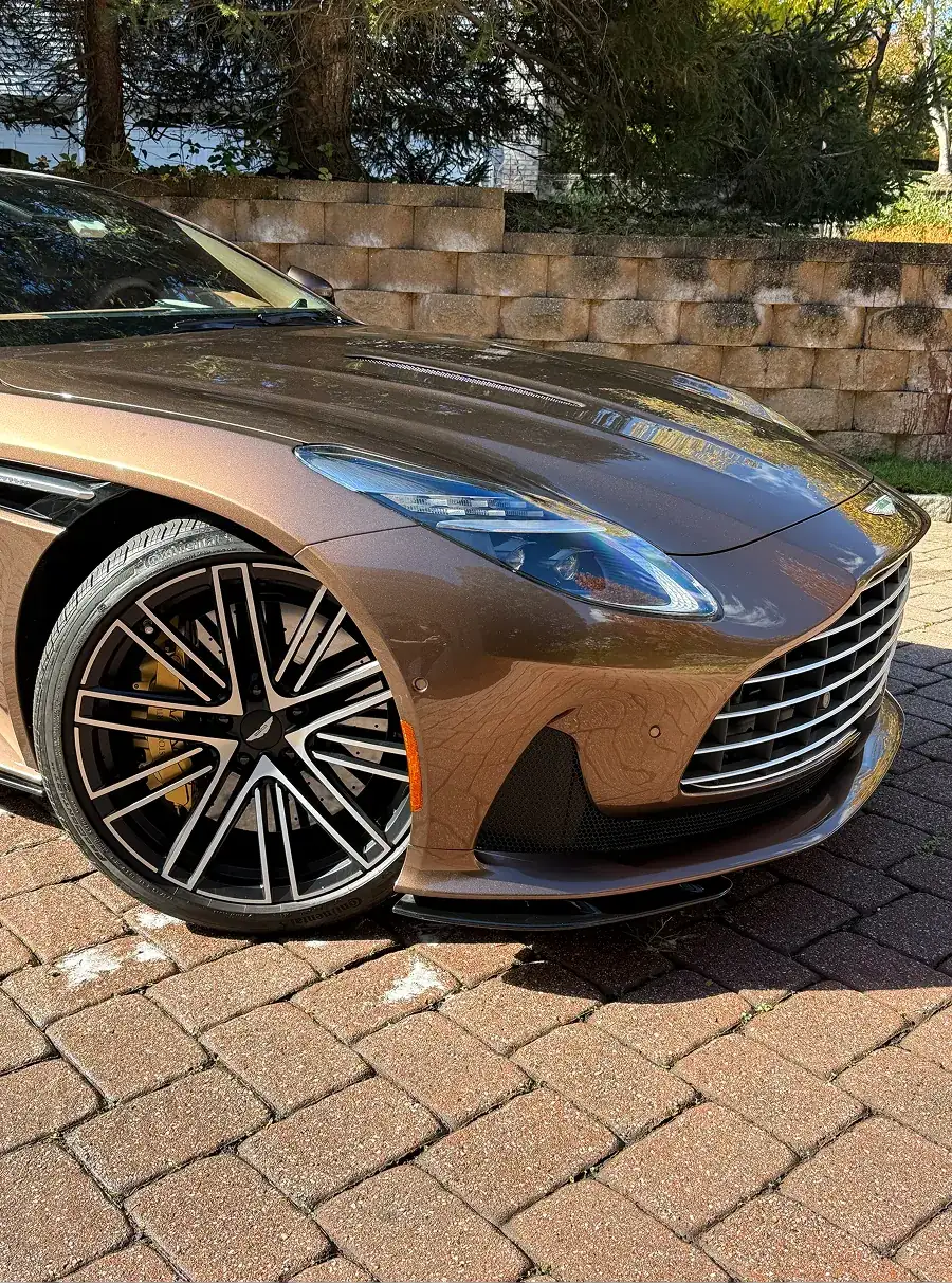 Front side of a brown luxury sports car with detailed wheel and distinctive grille parked on a brick driveway.