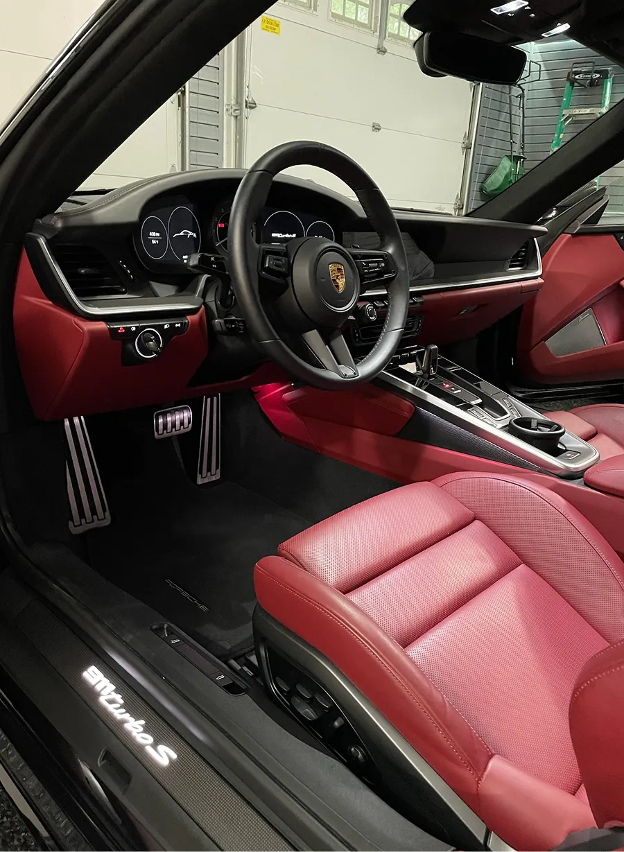 Interior of a Porsche 911 Turbo S showing the black steering wheel with Porsche emblem, red leather seats, and pedal area.