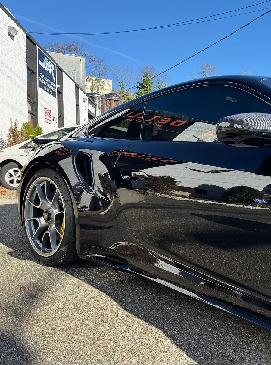 Close-up side view of a black sports car parked on asphalt with buildings and blue sky in the background.
