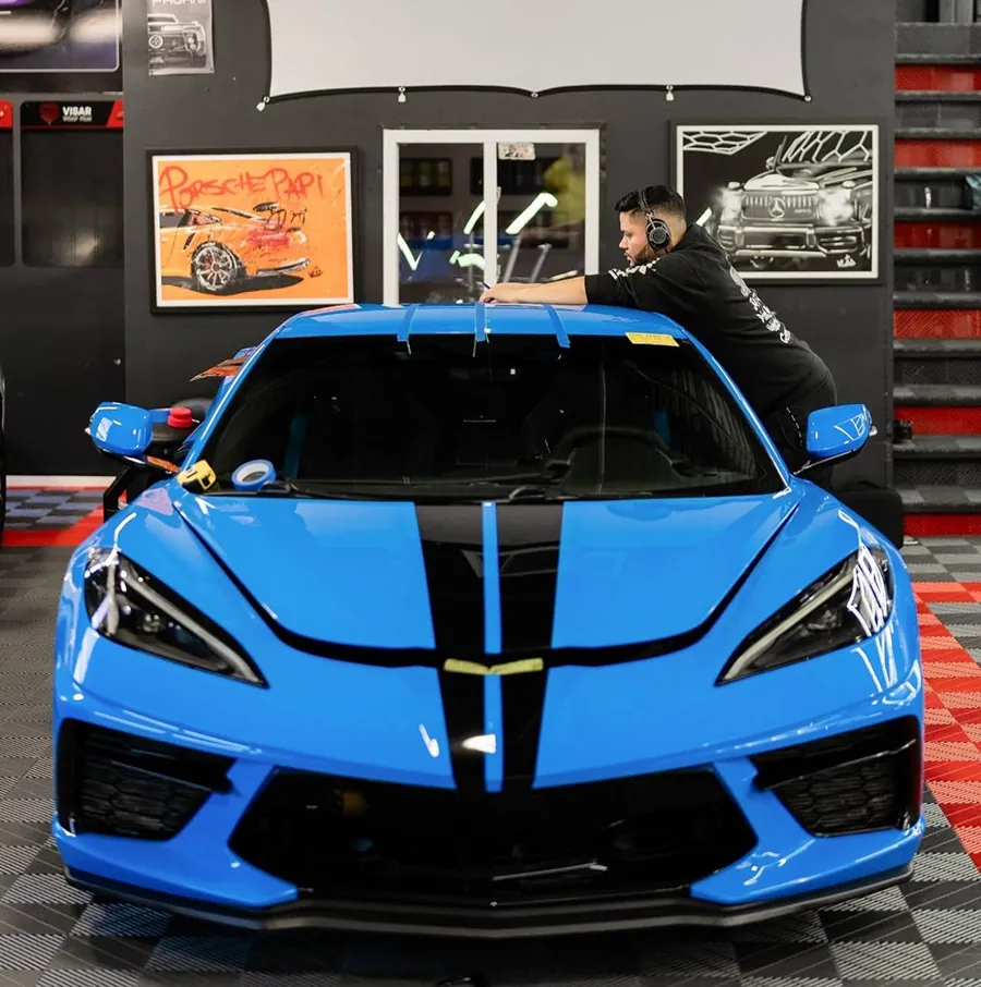 Blue sports car with black racing stripes parked indoors, with a man wearing headphones leaning on its roof.