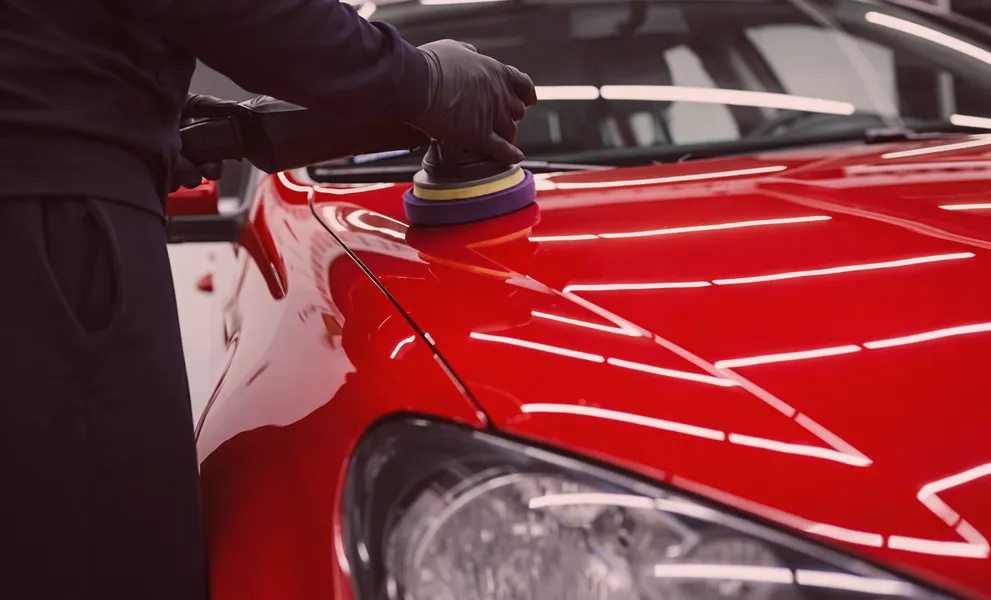 Person wearing black gloves polishing the shiny red hood of a car with a buffer machine.