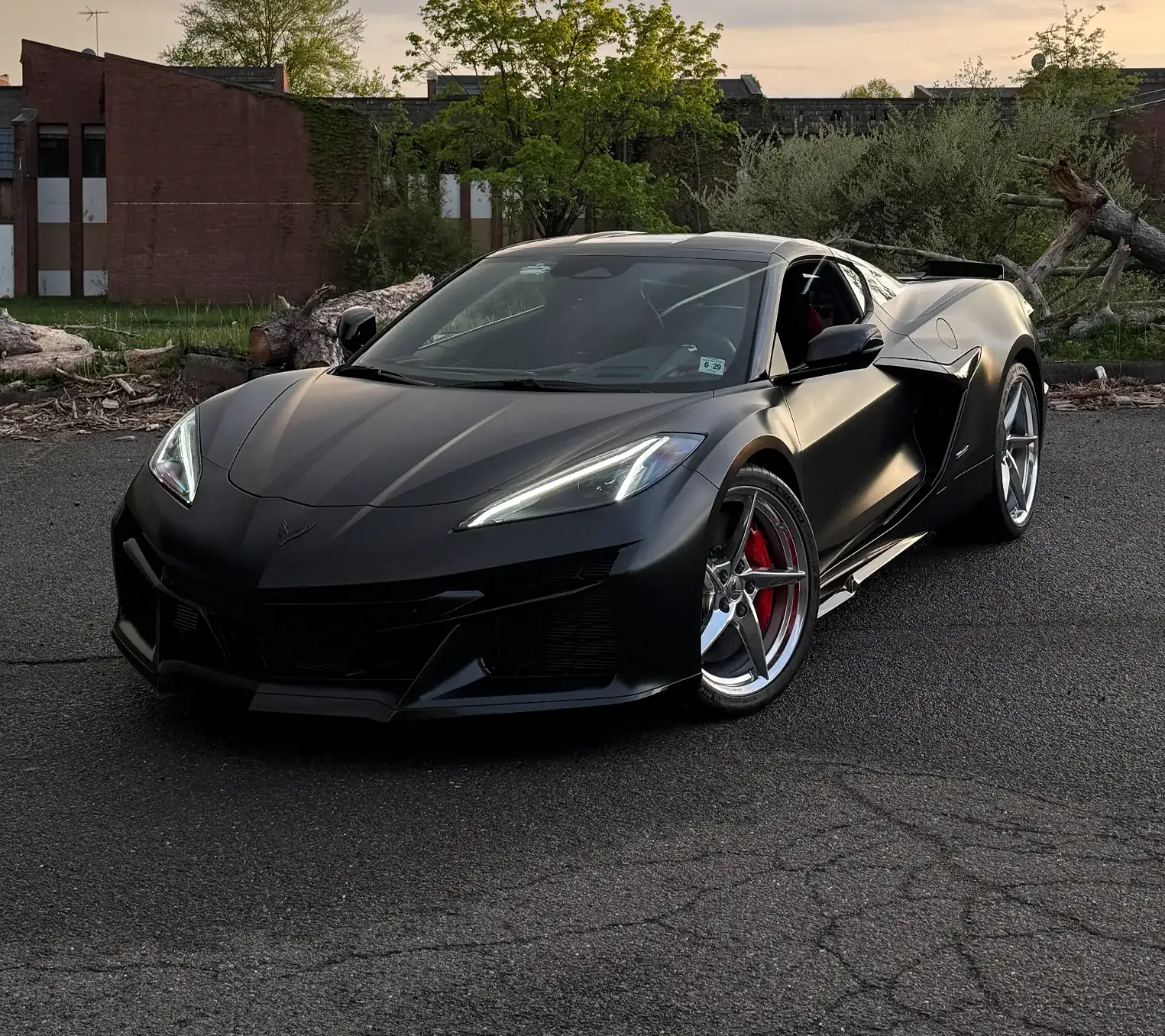 Matte black Chevrolet Corvette sports car parked on asphalt with trees and a brick building in the background.