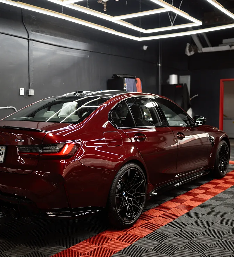 Shiny dark red BMW M3 sedan parked on a black and red checkered floor inside a garage with bright ceiling lights.