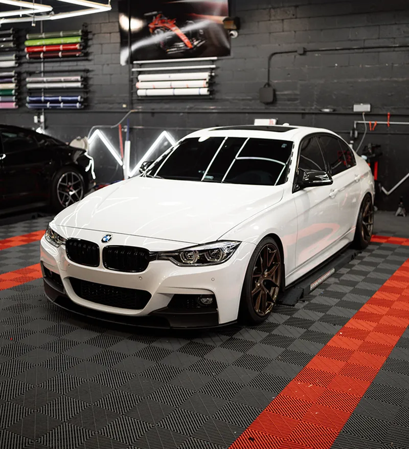 White BMW sedan with dark bronze wheels parked inside a garage with patterned flooring and racing posters on the wall.