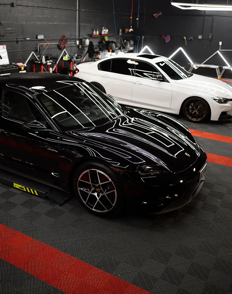 Black Porsche sports car and white BMW sedan parked side by side in a garage with black and red checkered flooring.