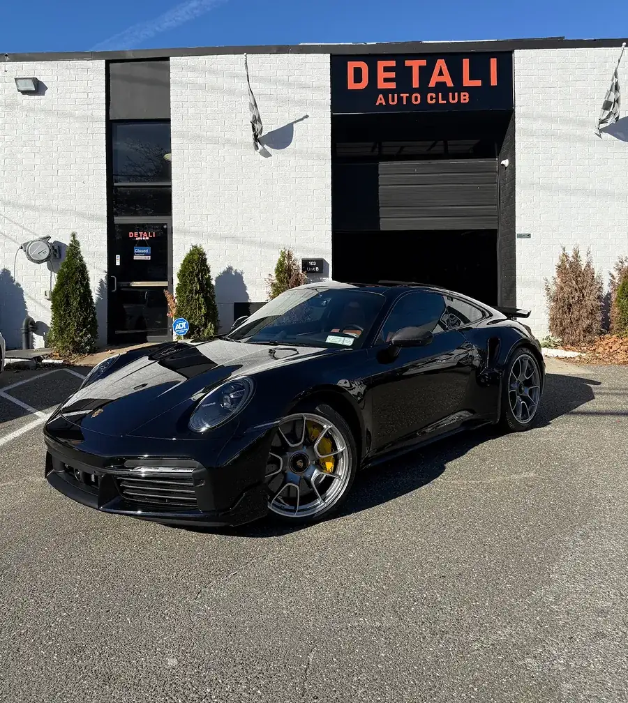Black Porsche sports car parked in front of a white building with a sign reading 'DETALI AUTO CLUB'.