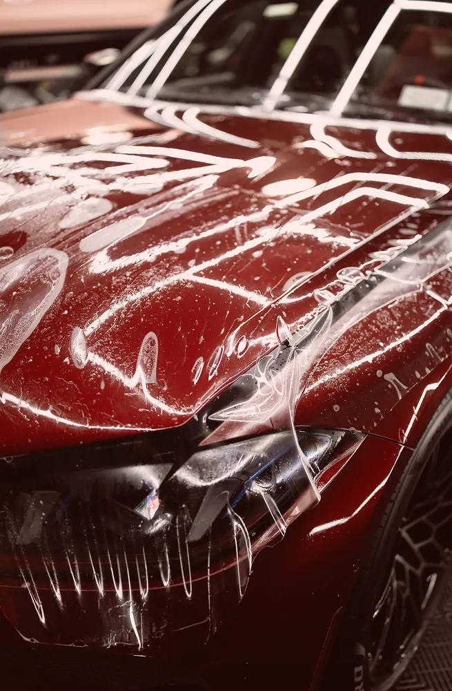 Close-up of a red car front with a protective clear film being applied over the headlight and hood.