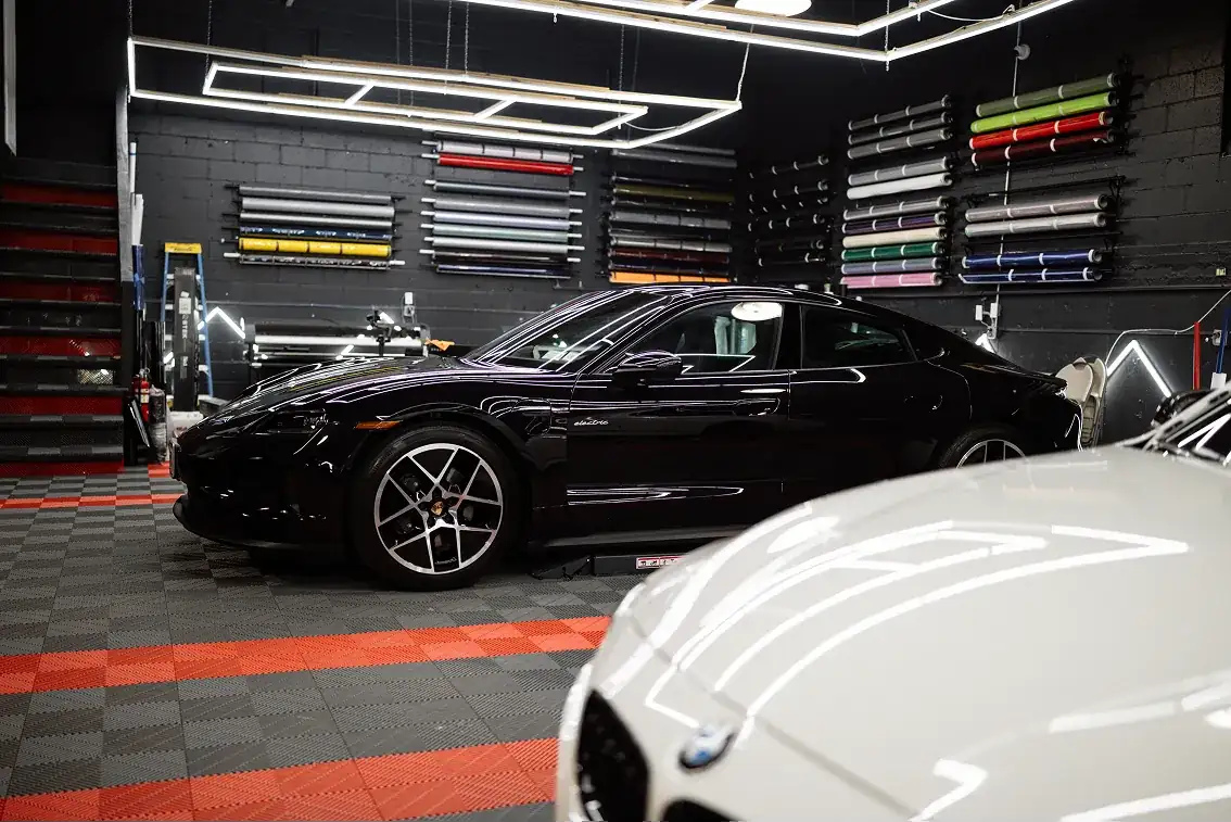 Black Porsche Taycan parked inside a garage with various colored vinyl wrap rolls mounted on the wall.