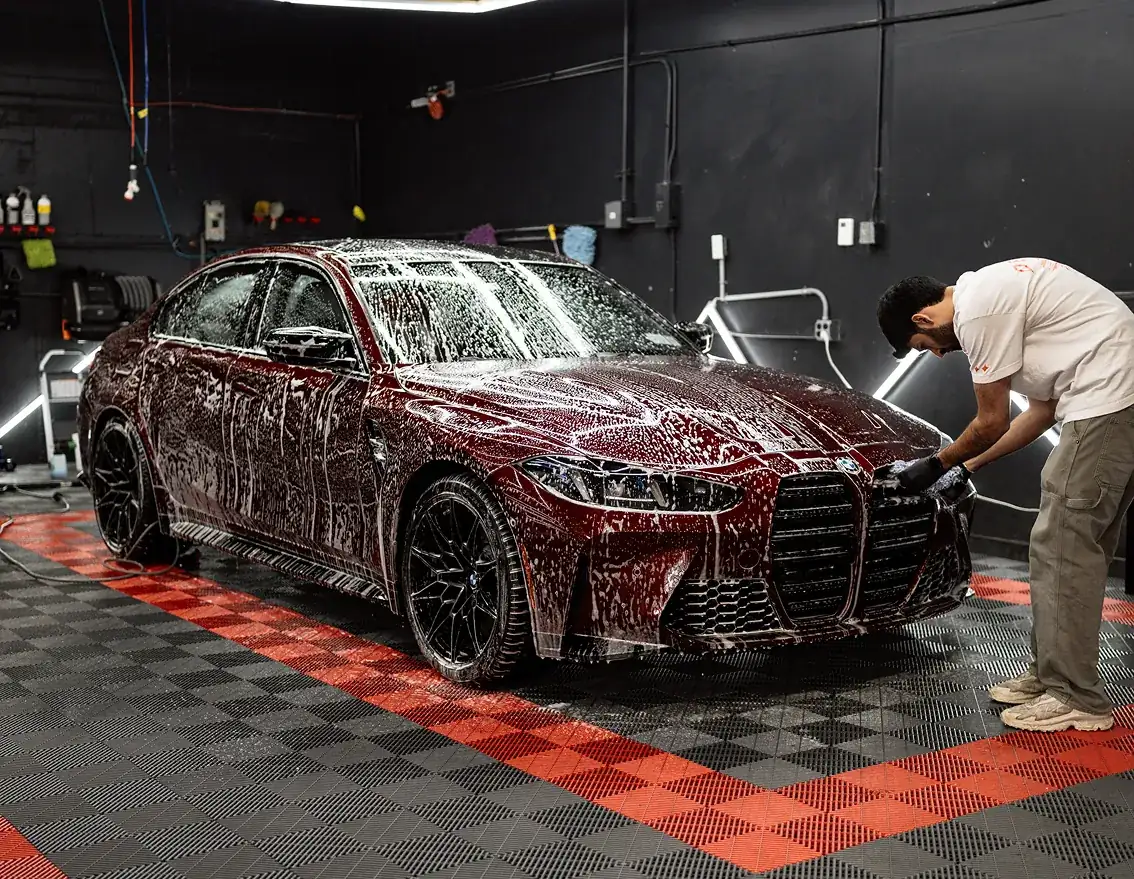 Man washing a dark red BMW covered in soap foam inside a garage with black and red checkered flooring.