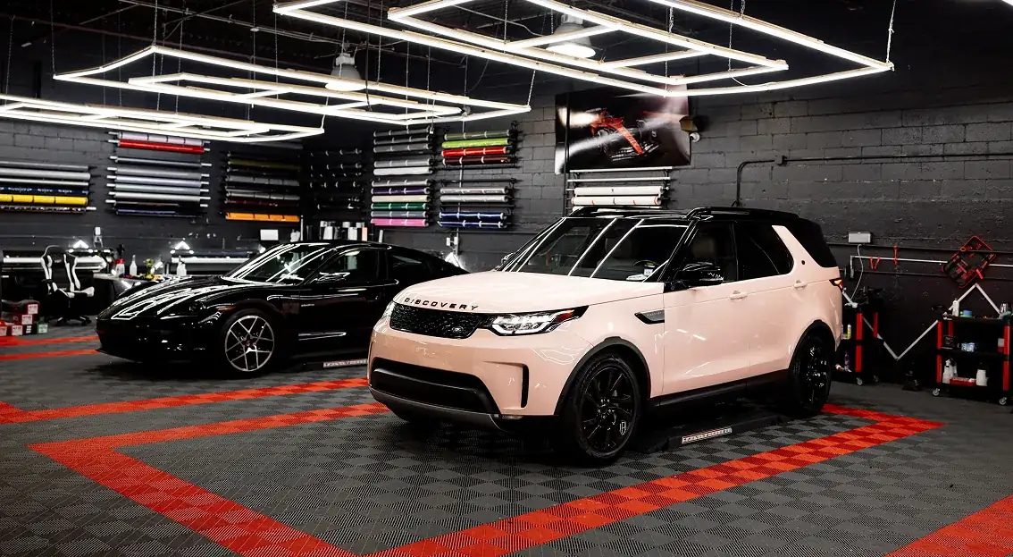 A white Land Rover Discovery and a black sports car parked inside a modern garage with bright rectangular ceiling lights and colorful wrap rolls on the wall.