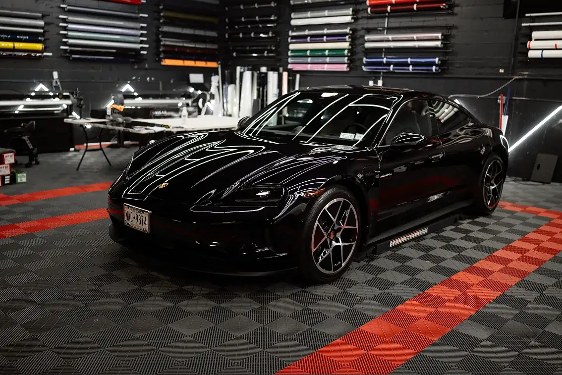 Black Porsche Taycan electric car parked in a workshop with wrap rolls on the wall and checkered flooring with red stripes.