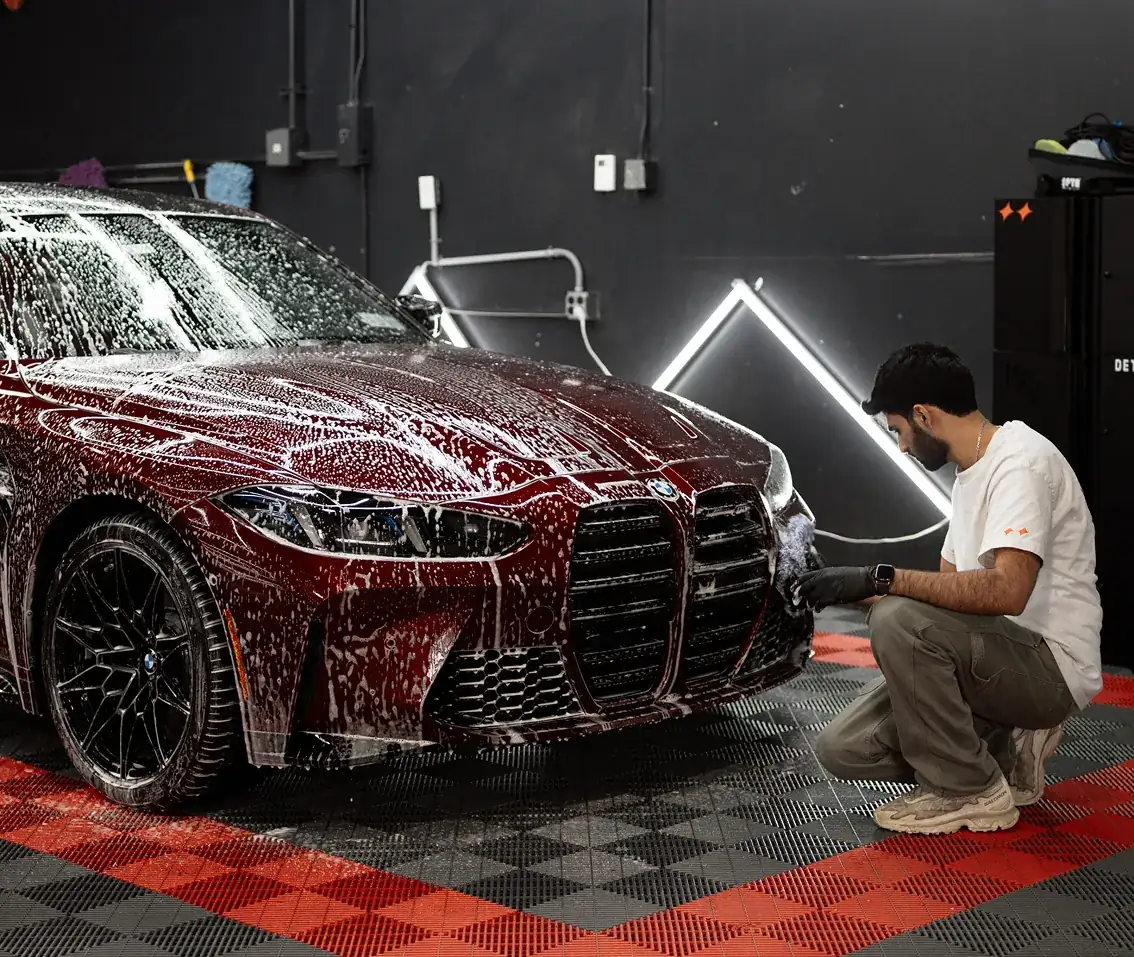 Man wearing gloves washing the front of a red BMW covered in soap suds inside a garage with black walls and red and gray checkered flooring.