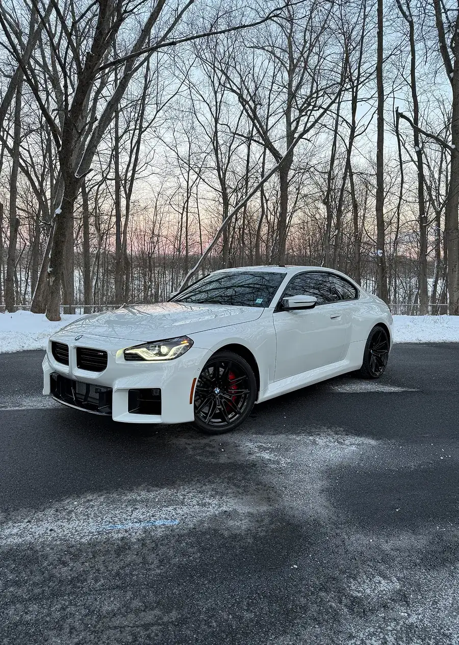 White BMW coupe parked on icy asphalt with bare trees and a sunset sky in the background.