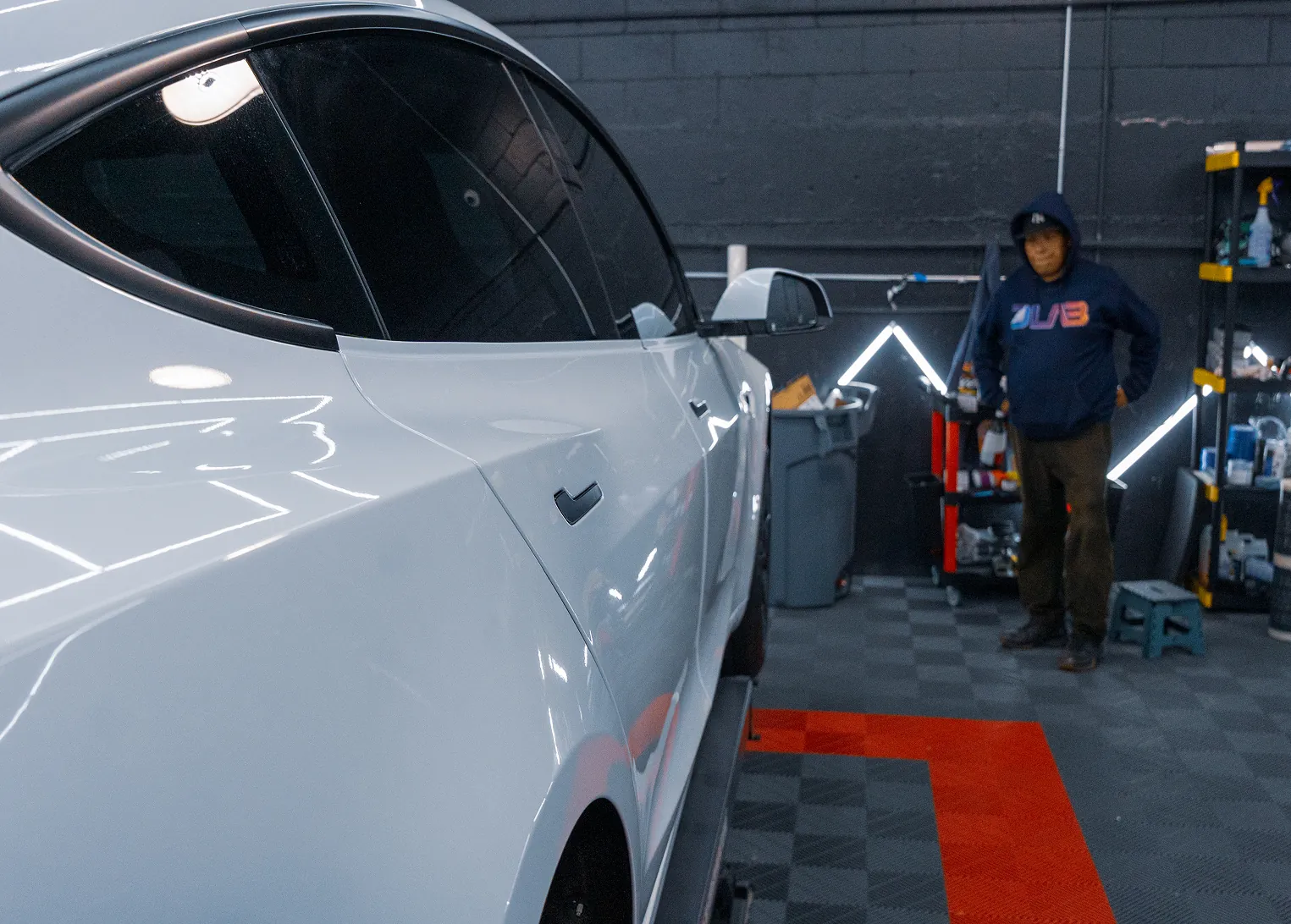 Person in a dark hoodie standing in a garage near a white car with reflective windows.