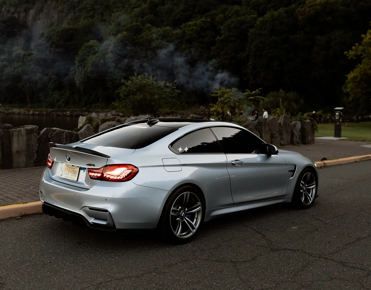 Silver BMW M4 coupe parked on a road near stone barriers and a wooded park area at dusk.
