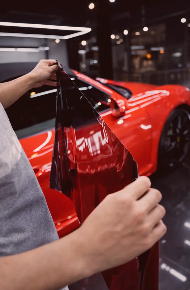 Person holding dark tinted film near a red car inside a garage or workshop.