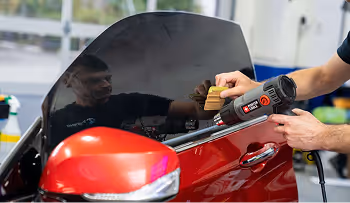 Person holding a heat gun and a brush near the side mirror of a red car with a tinted window.