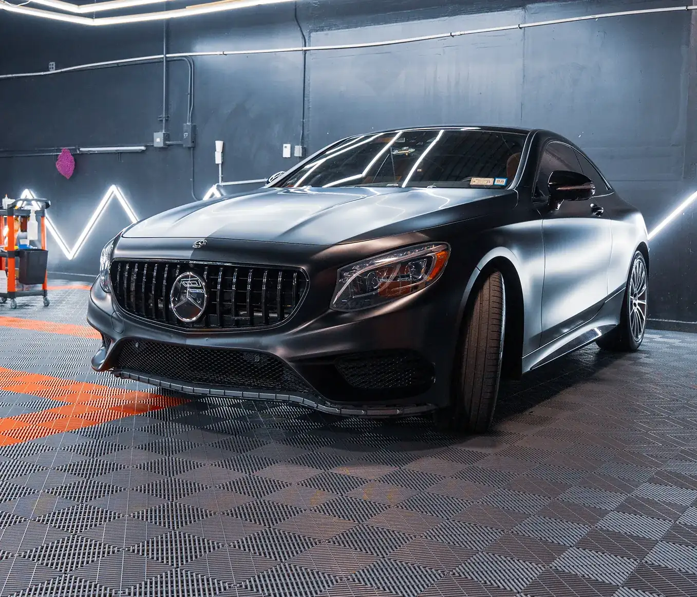 Front side view of a matte black Mercedes-Benz car parked indoors with modern lighting on the walls.