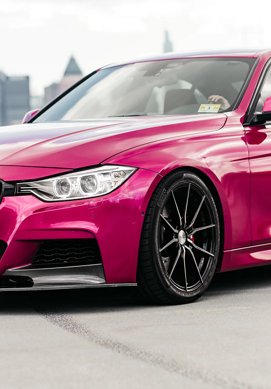 Front side view of a shiny pink sports car with black rims parked on a rooftop with city skyline in the background.