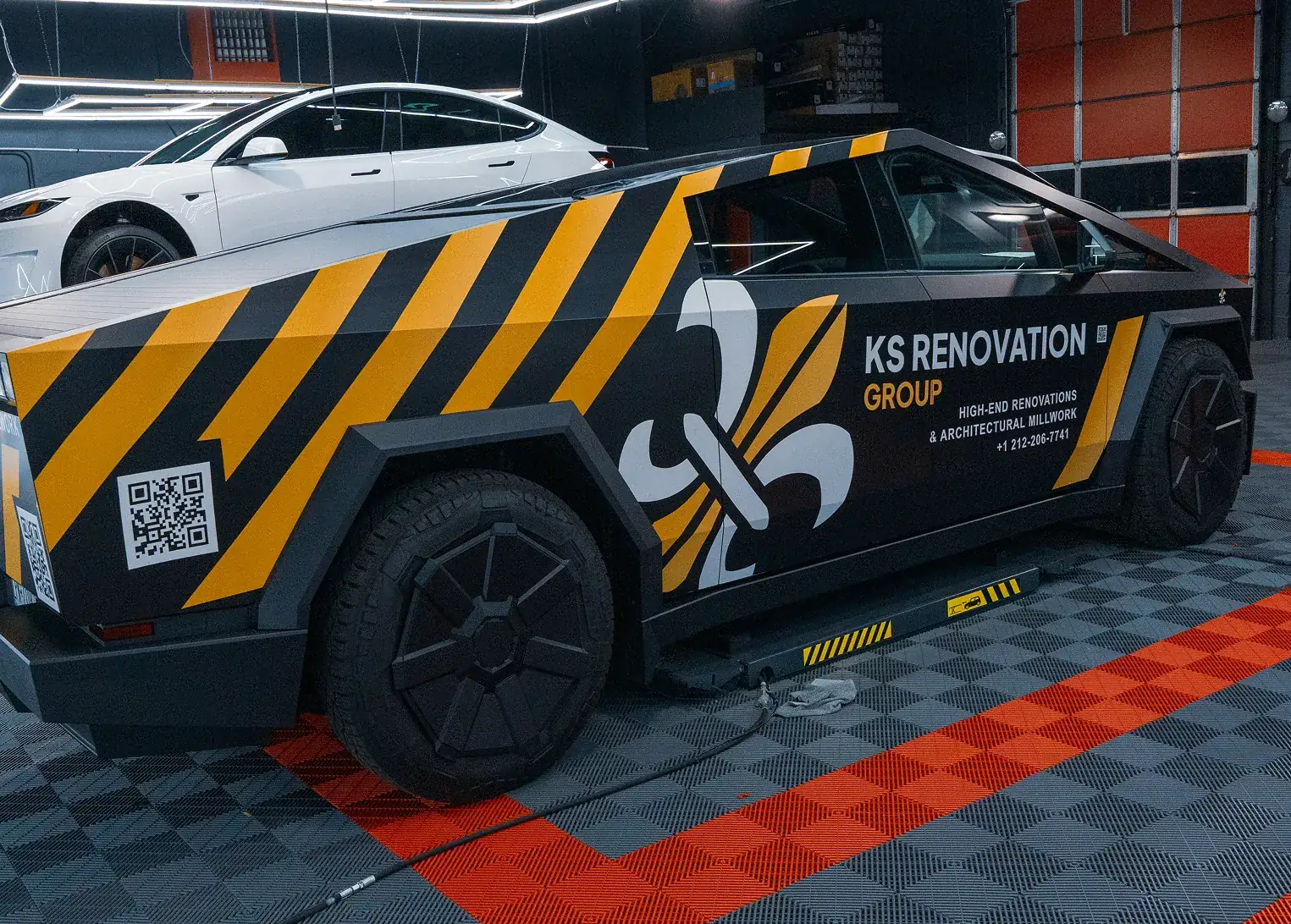 Black and yellow striped Tesla Cybertruck with KS Renovation Group branding and QR code parked inside a garage with a white Tesla Model 3 in the background.