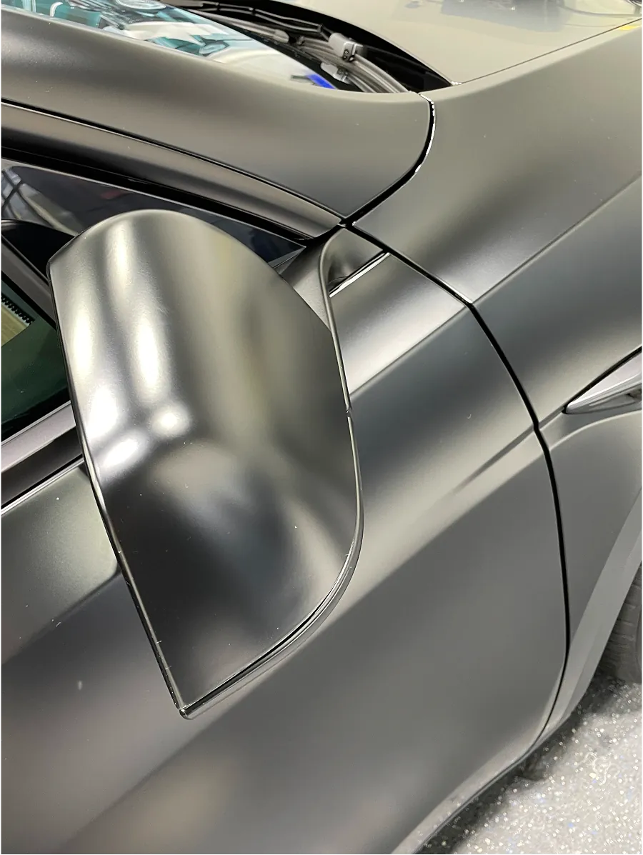 Close-up of a matte black car's side mirror and door panel with visible body lines and reflections.