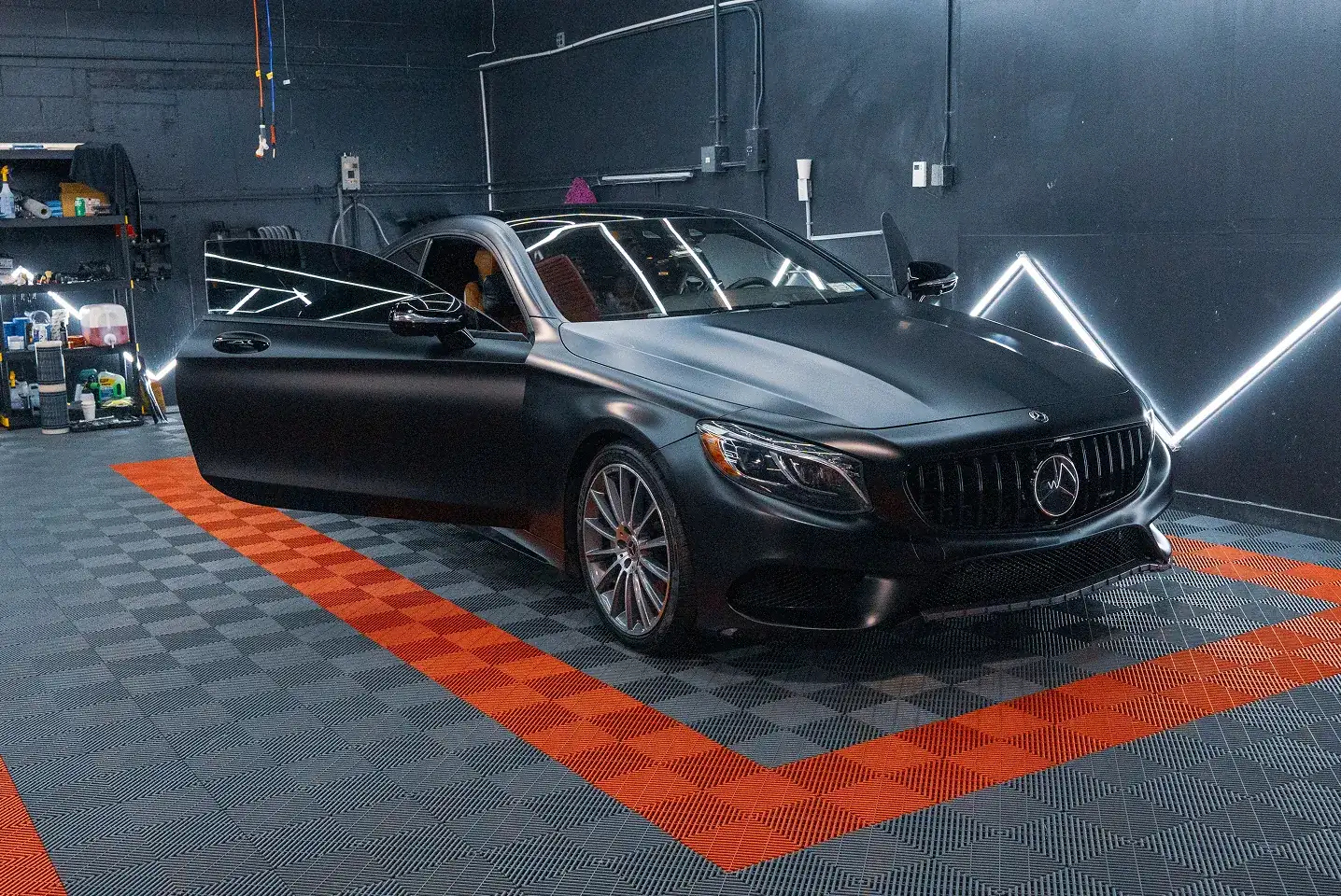 Black luxury sedan with front driver-side door open, parked on checkered floor with red border in a garage with dark walls and neon lights.