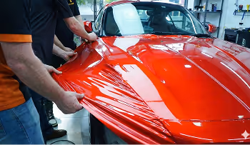 Two people applying a large sheet of red vinyl wrap onto the hood of a red car inside a workshop.